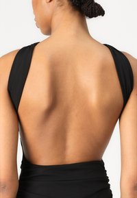 Melnā peldkostīmā ar dziļu izgriezumu mugurā, platām siksnām un savāktu audumu jostasvietā. Gluda tekstūra izceļ pieguļošu dizainu.