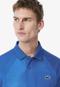 Blå polo skjorta i ett strukturerat, andningsbart material, med en tvåknappsslutning och en liten broderad logotyp på vänster bröst.