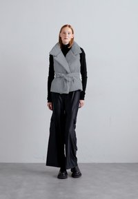 Grå pufferväst med bred krage och bälte i midjan, kombinerad med svarta vida byxor. Slät textur med en minimalistisk design.