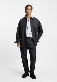 Uomo che indossa una giacca di denim scuro e jeans con una camicia a righe verticali bianche e nere e scarpe nere, in piedi contro uno sfondo bianco.