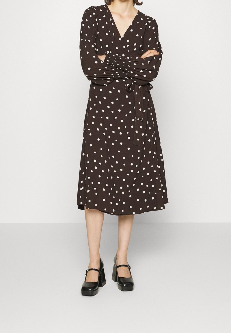 Robe marron à pois avec un décolleté en V, des manches longues et une taille nouée. Présente un design fluide et tombe en dessous du genou. Chaussures à plateforme noires.