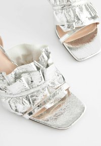 Sølvmetallic sandaler med en tekstureret overdel med rynkede detaljer og firkantet tådesign. Åben hæl og flad sål.