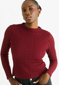 Burgundy long-sleeve top with textured fabric featuring a subtle pattern, crew neckline, and fitted design. Paired with dark jeans.