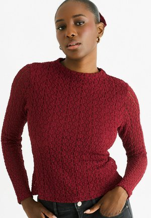 Burgundy long-sleeve top with textured fabric featuring a subtle pattern, crew neckline, and fitted design. Paired with dark jeans.