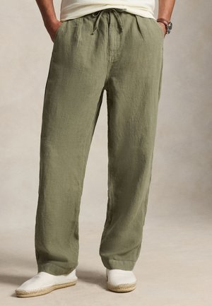 Homme portant un pantalon large en lin vert olive avec une taille élastique et des espadrilles blanches à enfiler, mains dans les poches.