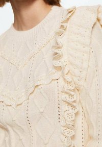 Cremefarbener gestrickter Pullover mit Zopfmuster und Rüschenakzenten entlang der Schulter und des Ausschnitts. Die Textur wirkt weich.