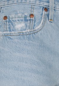 Jeans in denim blu chiaro con una tasca posteriore consumata e bordi sfilacciati, rivetti in rame e cuciture verticali lungo la vita.