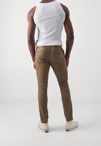 Tanské chinos s úzkým střihem, obsahující zadní kapsy, a nošené s bílými teniskami. Model nosí bílý top bez rukávů.