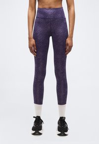 Personne portant des leggings violet à motifs taille haute, des chaussettes blanches courtes, et des chaussures de course noires et blanches, debout devant un fond uni.