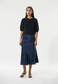 Schwarzer, gestrickter Kurzarmpullover mit strukturierten Punkten, kombiniert mit einem fließenden, marineblauen Satin-Midiglenk. Model trägt schwarze Riemchen-Heels.