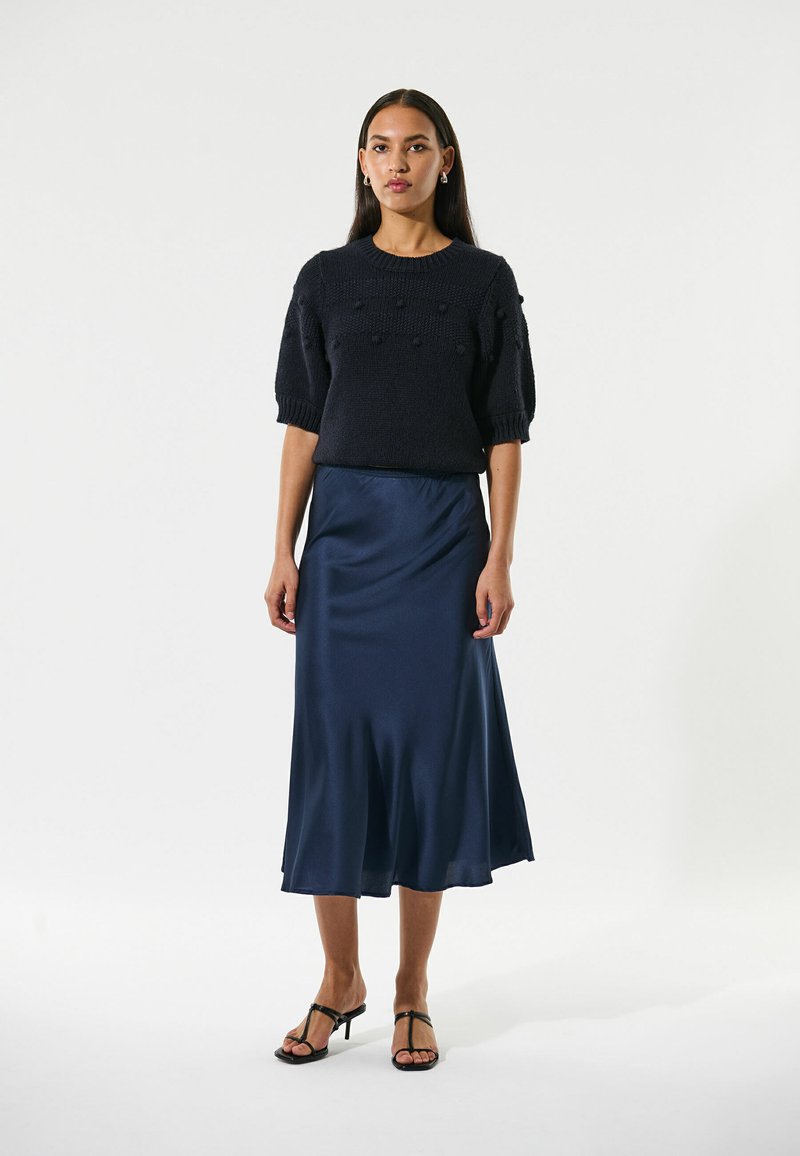 Schwarzer, gestrickter Kurzarmpullover mit strukturierten Punkten, kombiniert mit einem fließenden, marineblauen Satin-Midiglenk. Model trägt schwarze Riemchen-Heels.
