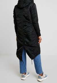 Czarna długa puchowa kurtka z kapturem, z dopasowanym ściągaczem u dołu i ściągaczami na rękawach, zestawiona z jasnoniebieskimi dżinsami i trampkami.