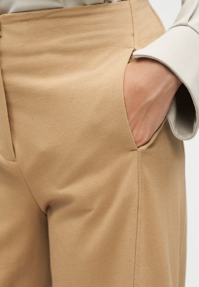 Χέρι που ακουμπά στην τσέπη μπεζ παντελονιού με διακριτικό σχέδιο ψαροκόκκαλου, φορώντας ανοιχτό μπεζ πουκάμισο με μακριά μανίκια.