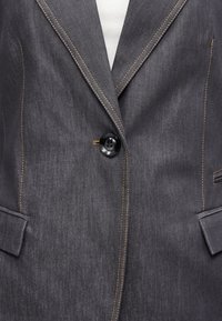 Σκούρο γκρί σακάκι από τζιν ύφασμα με πορτοκαλί ραφές, κλειστό με ένα μαύρο κουμπί, με τσέπες με καπάκι, φορεμένο πάνω από λευκή ριμπ ύφανση μπλούζας.