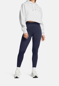 Skórzana szara bluza z kapturem z sznurkami, w zestawie z granatowymi legginsami. Model nosi białe sneakersy i skarpetki do kostek z prążkowanym ściągaczem.