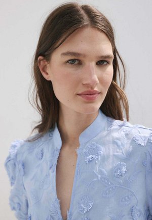 Joven mujer de piel clara y cabello castaño lleva una blusa azul claro bordada, mirando ligeramente fuera de cámara con expresión neutral.