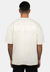 Weißes, übergroßes T-Shirt mit kurzen Ärmeln, auf dessen Rückseite das Wort "DROPSIZE" in fetter Schrift gedruckt ist. Aus weichem Baumwollmaterial.