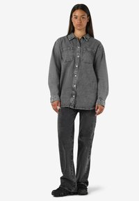 Üks suur suuruses hall denimist särk, millel on kaks esistikku, nööpidega ees ja langenud õlad, koos hallide denimipükste ja mustade kingadega.