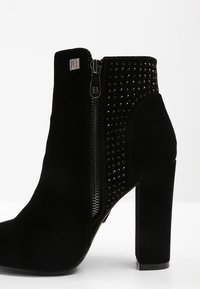 Černé semišové kotníkové boty na vysokém podpatku. Mají boční zip a ozdobné cvočky na jedné straně. Hladký povrch s elegantním designem.
