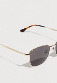 Gafas de sol Lacoste con montura dorada, lentes gris oscuro y terminales de varillas con diseño carey sobre una superficie blanca.