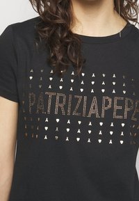 Camiseta de algodón negra con acentos en dorado metálico. En la parte frontal se muestra el texto "PATRIZIA PEPE" rodeado de formas decorativas de corazones y lazos.