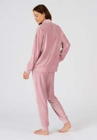 Conjunto de loungewear de terciopelo en rosa claro, que incluye una chaqueta con cremallera y un cuello alto, y pantalones cómodos con puños elásticos. Tejido suave y texturizado.