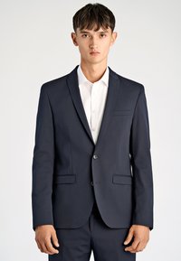 Marynarka w kolorze granatowym wykonana z gładkiej tkaniny, z dwoma przednimi kieszeniami, dwoma guzikami i klapą z wycięciem, w zestawieniu z białą koszulą na spodzie.