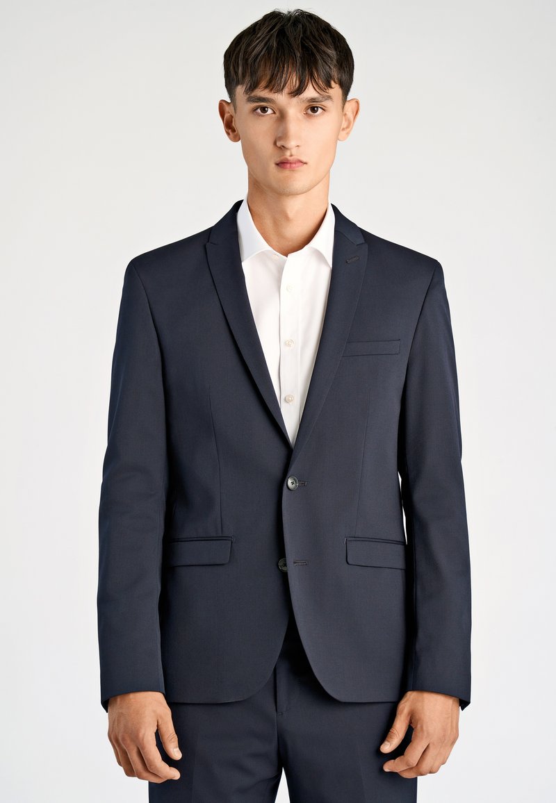 Marynarka w kolorze granatowym wykonana z gładkiej tkaniny, z dwoma przednimi kieszeniami, dwoma guzikami i klapą z wycięciem, w zestawieniu z białą koszulą na spodzie.