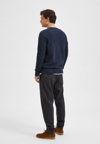 Dunkelblauer Strickpullover mit gerippten Bündchen, kombiniert mit dunkel gemusterten Hosen und braunen Wildlederstiefeln. Die Textur wirkt weich, der Schnitt lässig.