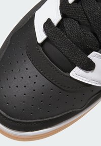 Svart sneaker med överdel i läder, vita accenter, perforerad yta, runda snören och sula i gummiliknande material. Har en rundad tådesign.