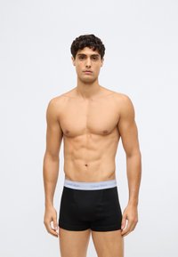 Jovem com cabelo curto e encaracolado de pé, de frente, usando boxers pretos da Calvin Klein com uma cinta cinza claro.