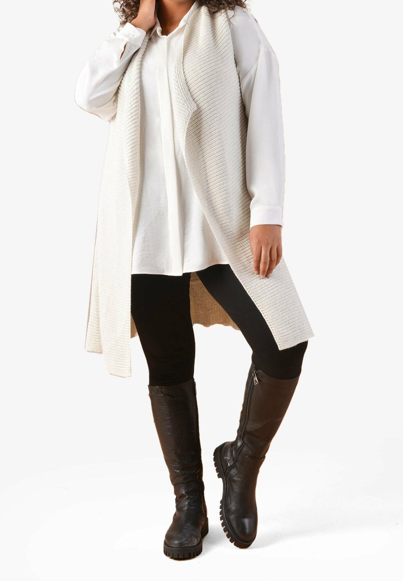 Long cream knit cardigan over a white shirt, black leggings, and knee-high black leather boots with a textured sole.
