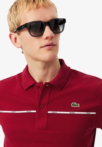 Blonde man met zwarte Lacoste zonnebril en een rode Lacoste polo met krokodillenlogo en witte merkstrepen over de borst.