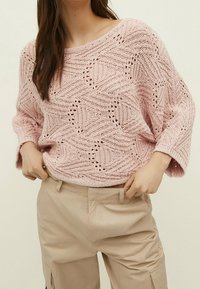 Frau trägt einen lockeren, hellrosa gestrickten Pullover mit geometrischen Mustern und beigen Hosen, während sie ihren Taillenbereich mit beiden Händen anpasst.