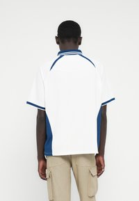 Λευκό πόλο πουκάμισο με ναυτικό μπλε λεπτομέρειες. Κοντά μανίκια, γιακάς με δύο ρίγες και υφασμένες πλευρικές πιέτες. Καθημερινός σχεδιασμός με άνετη εφαρμογή.