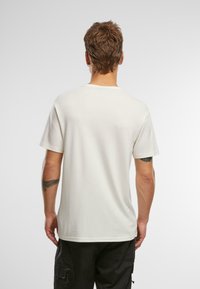 Krämfärgad bomulls-t-shirt med korta ärmar, rund hals, avslappnad passform och en enkel design. Visad från baksidan, med en synlig tatuering på armen.