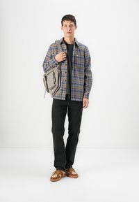 Plaid button-up shirt in blue and yellow, black t-shirt, black jeans, and brown sneakers with a textured upper; carrying a grey shoulder bag.