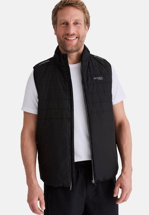 Lächelnder Mann trägt eine schwarze gesteppte ärmellose Weste über einem weißen T-Shirt mit kurzen Ärmeln und steht vor einem schlichten weißen Hintergrund.