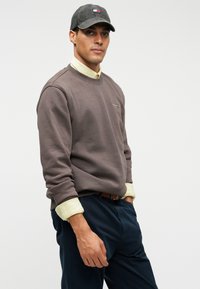 Uomo che indossa un berretto grigio, una felpa marrone sopra una maglietta gialla e pantaloni blu, in piedi con una mano in tasca su uno sfondo semplice.