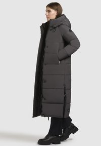 Cappotto puffer lungo nero con cappuccio, bottoni a pressione e zip laterali. Realizzato in un materiale liscio e isolante con una vestibilità oversize.