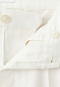 Gros plan sur la ceinture d'un pantalon blanc cassé montrant un bouton, une boutonnière, une fermeture éclair et une bande élastique intérieure réglable avec bouton.