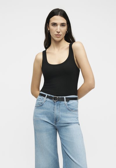 Νέα γυναίκα με μακριά σκούρα μαλλιά, φορώντας μαύρο τοπ μπλούζα στενωμένη μέσα σε ανοιχτό μπλε ψηλόμεσο τζιν με μαύρη ζώνη, στέκεται με τα χέρια πίσω από την πλάτη.