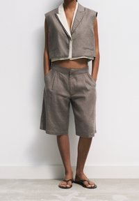 Persona vestida con un chaleco gris sin mangas y corto, combinado con pantalones cortos de tiro alto que llegan hasta las rodillas y sandalias de playa negras, de pie contra una pared blanca.