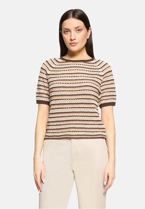 Femme aux cheveux bruns lisses portant un haut en maille à manches courtes rayé marron et beige, et un pantalon beige clair, debout devant un fond uni.