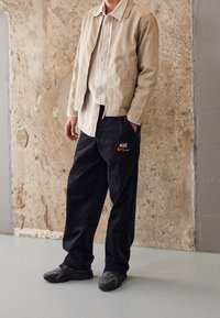 Beige bomullskavaj över en ljus skjorta med knappar, kombinerad med svarta Nike-byxor med logotyp och grå sportskor med mörka accenter.