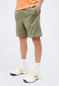 Άτομο που φοράει σορτς ελιάς με το λογότυπο Lacoste, λευκές κάλτσες μέχρι τους αστραγάλους και λευκά αθλητικά παπούτσια Lacoste, στέκεται με τα χέρια στις τσέπες.