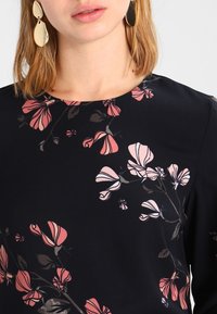 Top in tessuto nero con motivi floreali rosa, scollatura rotonda e texture liscia. Orecchini statement dorati completano il design.