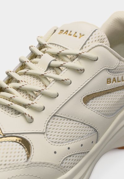 Κοντινό πλάνο μπεζ αθλητικού παπουτσιού Bally με δίχτυ, χρυσές λεπτομέρειες και υφασμένα κορδόνια περασμένα από οπές.