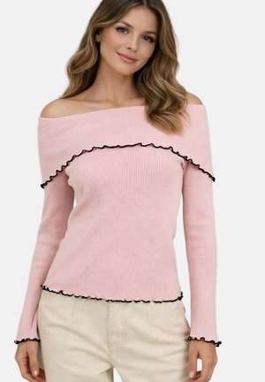Femme portant un pull rose côtelé à épaules dénudées avec des bords noirs à volants et un pantalon beige, souriante avec des cheveux ondulés détachés.