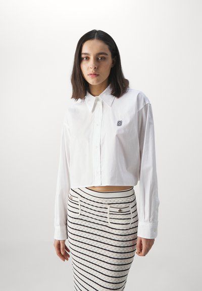 White cropped shirt with long sleeves and button front, featuring a small black logo. Paired with a textured, striped pencil skirt.